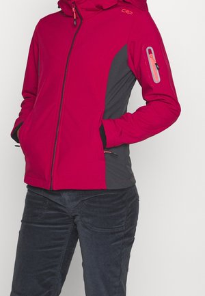 Kvinde iført en rød og grå hættetrøje med lynlåslommer i siderne og en lynlåslomme på den øverste venstre ærme, kombineret med mørke bukser.