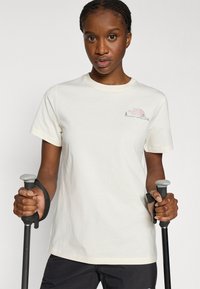 Ανοιχτόχρωμη κρεμ βαμβακερή κοντομάνικη μπλούζα με στρογγυλή λαιμόκοψη, κοντά μανίκια και μικρό ροζ λογότυπο στο αριστερό στήθος. Άτομο κρατάει μαύρα μπαστούνια πεζοπορίας.