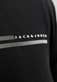 Black t-shirt featuring silver-textured "JACK & JONES" logo and horizontal lines. Material appears smooth with a fitted design.