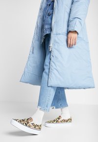 Giacca imbottita azzurro chiaro, jeans in denim e scarpe slip-on con stampa leopardata multicolore e suola bianca, abbinate a calze grigie chiare.