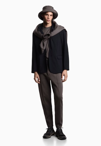 Jonge man met een taupe bucket hat, een taupe sweatshirt over een zwarte blazer, taupe broek en zwarte sneakers op een witte achtergrond.