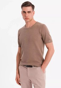 Top de punto beige de manga corta con un patrón texturizado, escote redondo y corte ajustado. Combinado con pantalones de color claro y un cinturón.