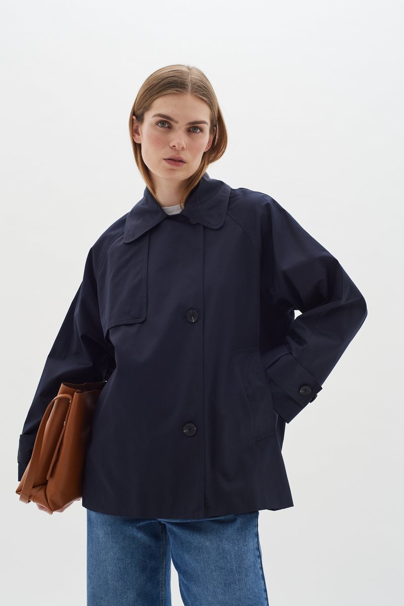 Tumši zils, ūdensizturīgs jakas ar lielu apkakli, priekšējiem kabatām un pogu aizdari. Modelis tur brūnu somu, kombinēts ar džinsu biksēm.