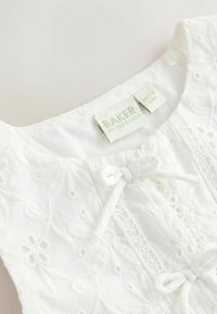 Encolure en tissu blanc brodé avec bouton et liens, avec étiquette de vêtement "Baker by Ted Baker" sur un matériau doux.