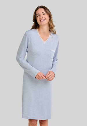 Mujer sonriente que lleva un vestido de manga larga hasta la rodilla de color azul claro, con detalles de encaje en el escote en V y un bolsillo en el pecho, de pie contra un fondo gris.