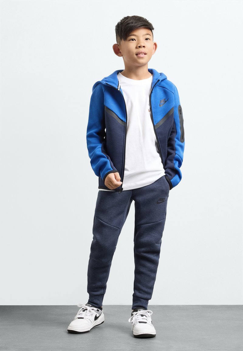 Set di abbigliamento sportivo Nike composto da un giubbino blu e blu navy con zip, una T-shirt bianca, pantaloni jogger blu navy e sneakers bianche con dettagli neri.