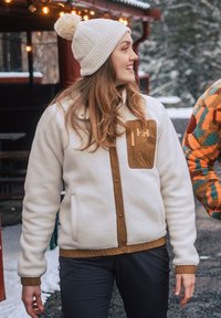 Kreemikas fleece-jakk, millel on pruun tasku, nööbid ja mansetid. Kandmisel kreemivärvi kootud beanie pom-pom detailiga.