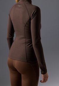 Top deportivo de manga larga marrón con cuello alto, diseño ajustado, bordes sin costuras y textura suave, combinado con leggings marrones a juego.