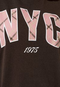 T-shirt marrone con lettere oversize a quadri bianchi e rosa che scrivono "NYC" e "1975" in bianco, con una texture di cotone liscia.