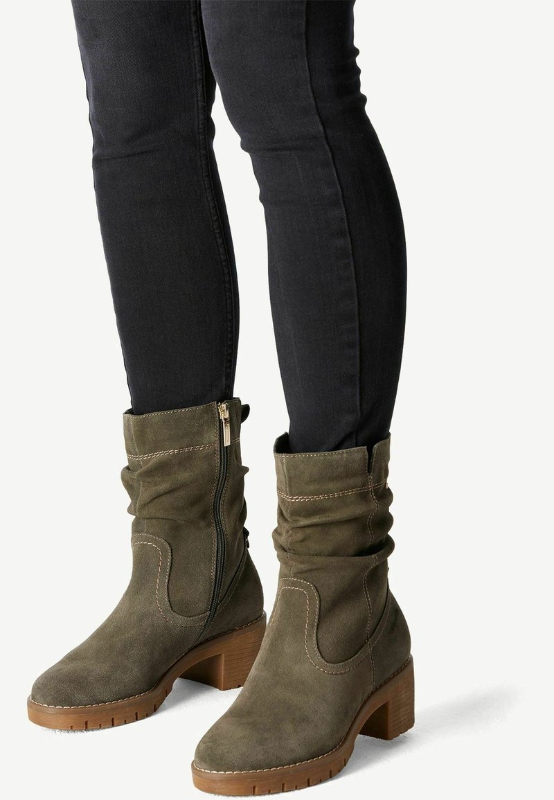 Personne portant des bottes mi-mollet en daim vert olive avec des talons en bois épais et des fermetures éclair latérales, associées à un pantalon noir ajusté.