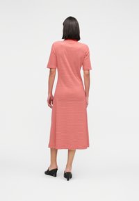 Γυναίκα που στέκεται, φοράει midi φόρεμα με κόκκινες και λευκές ρίγες, κοντά μανίκια και μαύρα mule με τακούνι, εμφανιζόμενη από πίσω.