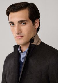 Cappotto di lana scuro con colletto alto che presenta un tessuto marrone a contrasto. Sotto, si vedono una camicia azzurra chiara e un maglione scuro.