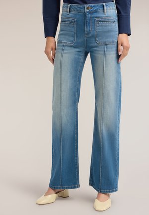 Kvinde iført lyseblå jeans med vide ben og frontlommer samt cremefarvede sko med lave hæle, vist fra talje til fødder.