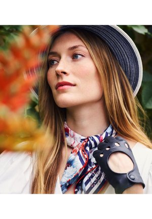 Frau mit langen blonden Haaren, die einen gestreiften Hut, einen floralen Schal und einen schwarzen fingerlosen Handschuh trägt und nachdenklich seitlich im Freien blickt.