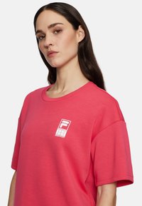 Lyserød oversized t-shirt lavet af bomuldsblanding, med et hvidt logo på brystet. Korte ærmer og rund halsudskæring. Glat tekstur.