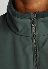 Giacca verde scuro a colletto alto, caratterizzata da una cerniera completa, una texture liscia e bordi rigati nel colletto, che enfatizzano un design elegante.