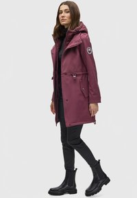 Maroon waterproof coat with a hood, drawstring waist, side pockets, and logo patch. Worn with black slim-fit pants and black ankle boots.
