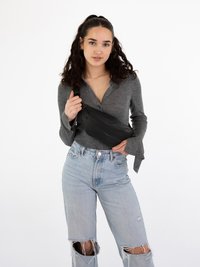 Schwarze Umhängetasche mit Reißverschluss, aus glattem Material, im Crossbody-Stil getragen. Modell in einem grauen Langarmoberteil und abgenutzten hellblauen Jeans.
