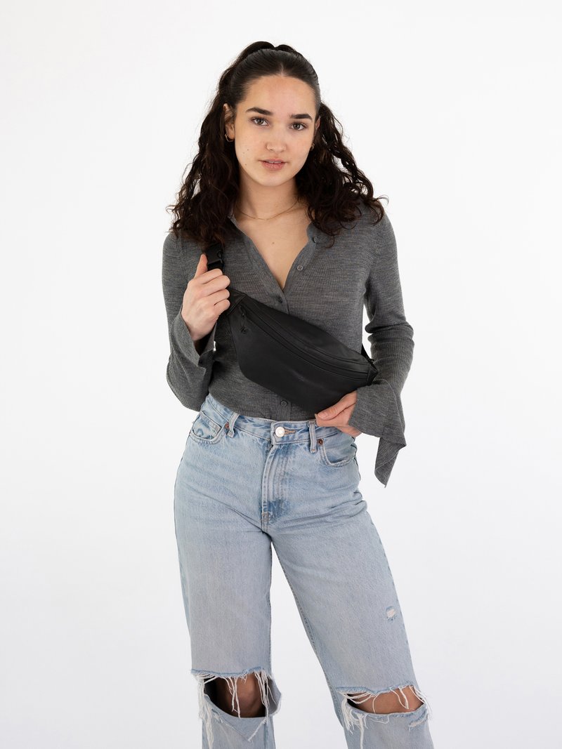 Marsupio nero con zip, realizzato in materiale liscio, indossato a tracolla. Modello con una maglia grigia a maniche lunghe e jeans chiari strappati.