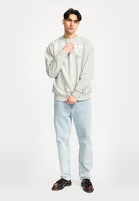 Lichtgrijs sweatshirt met geribbelde mouwen, voorzien van een bedrukt logo en tekst; gecombineerd met lichtblauwe jeans en donkere schoenen. Effen achtergrond.