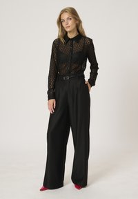 Sheer black lace button-up shirt with a collar over a black camisole; paired with high-waisted black wide-leg trousers and bright red heels.