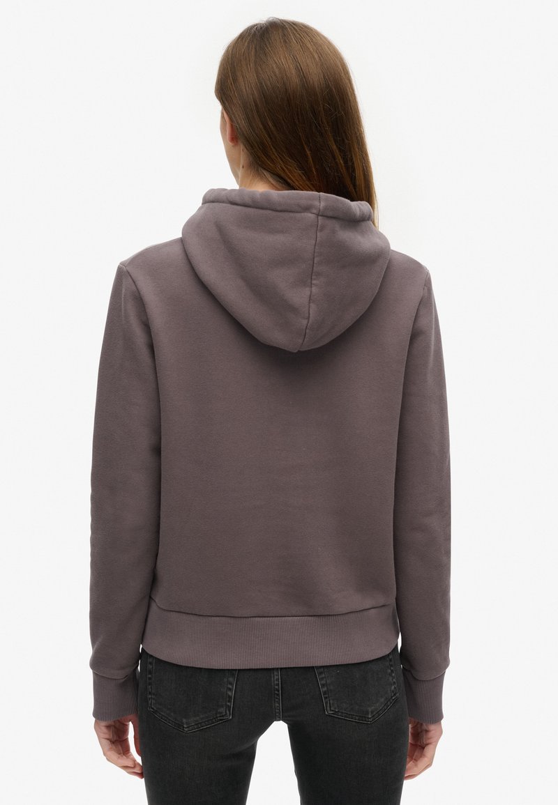 Kobieta z długimi brązowymi włosami ubrana w stonowaną fioletową bluzę z kapturem i czarne dżinsy, stojąca plecami do białego tła.