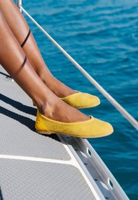 Žluté baleríny z filcu s hladkým povrchem, kulatou špičkou a malým akcentem poblíž paty, umístěné na texturovaném šedém povrchu.