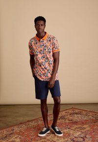 Korte mouw polo shirt in perzik met een bloemenprint, oranje kraag, gecombineerd met een marineblauwe korte broek en marineblauwe sneakers. Gelaagde, casual stijl.