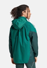 Giacca leggera verde e verde scuro con cappuccio, polsini elasticizzati e un design semplice. Mostra una trama liscia e accenti minimi.