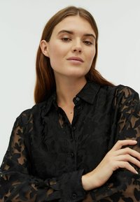 Bluză neagră transparentă cu model floral, închidere cu butoane în față și guler. Mâneci lungi cu mansete cu butoane și țesătură texturată.