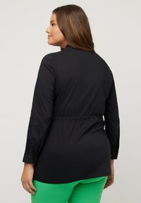Donna con lunghi capelli castani, che indossa una maglietta nera a maniche lunghe e pantaloni verde brillante, rivolta leggermente a sinistra su uno sfondo grigio uniforme.