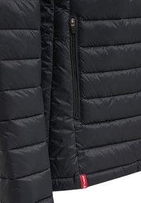 Giacca a vento in un blu scuro con motivo trapuntato, con tasca laterale con zip e un'etichetta logo rossa in basso. Texture liscia e brillante.