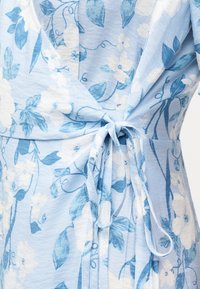 Φόρεμα τύπου wrap σε ανοιχτό μπλε χρώμα με λευκό και πιο σκούρο μπλε λουλουδένιο σχέδιο, δεμένο με φιόγκο στη μέση.