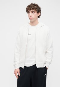 Λευκό φούτερ με φερμουάρ, με απαλή υφή, διαθέτει δύο μπροστινές τσέπες και κουκούλα με κορδόνι, φορεμένο πάνω από λευκό t-shirt με μαύρο κείμενο.