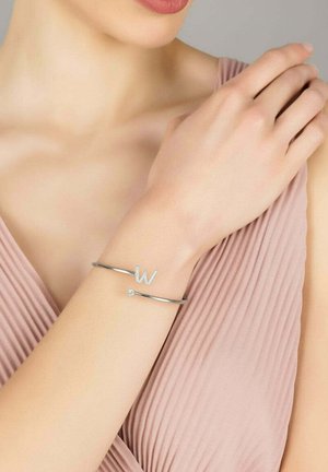 Silver bracelet featuring a capital "W" accent and a small diamond-like detail, set against a light pink fabric background.