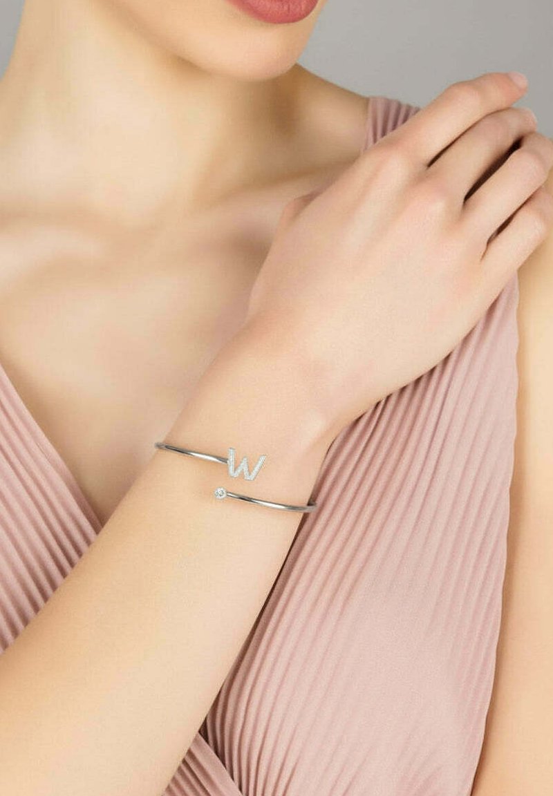 Silver bracelet featuring a capital "W" accent and a small diamond-like detail, set against a light pink fabric background.