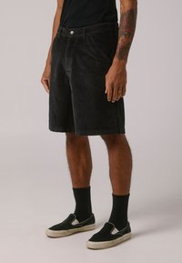 Schwarze Cordshorts mit geradem Schnitt, ausgestattet mit Fronttaschen, einem einzelnen Knopfverschluss und einem strukturierten Stoff. Kombiniert mit schwarzen Socken und Slippern.