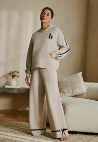 Helebeeži fleece-tüüpi kapuutsiga tõmblukuga jakk musta logoga ja mustade detailidega, koos laia säärega ülikonnapükstega, millel on lipsud ääres.