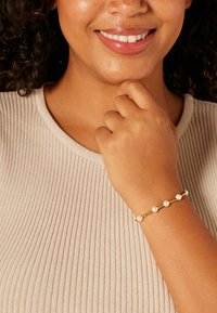 Gouden armband met zes kleine parels, gezet op een fijne ketting. De drager draagt een lichtbeige geribd shirt en rust haar hand nabij haar kin.