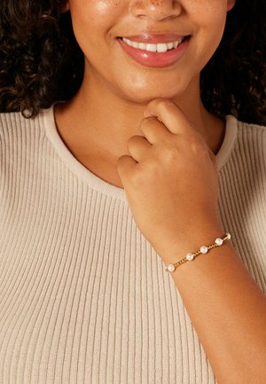 Gouden armband met zes kleine parels, gezet op een fijne ketting. De drager draagt een lichtbeige geribd shirt en rust haar hand nabij haar kin.