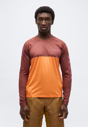 Hombre con una camiseta deportiva de manga larga en tonos naranja y marrón con el logo "ENDURA", de pie frente a un fondo claro y liso.
