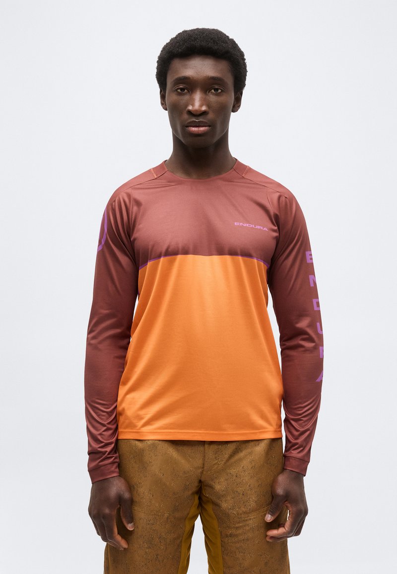 Άνδρας που φοράει αθλητική μπλούζα με μακριά μανίκια, δίχρωμη σε αποχρώσεις πορτοκαλί και καφέ με το λογότυπο "ENDURA", στέκεται μπροστά σε απλό ανοιχτόχρωμο φόντο.