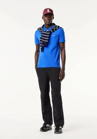 Camisa polo azul con un pequeño logo, pantalones cargo negros, suéter a rayas atado alrededor de los hombros y una gorra burdeos con un logo.