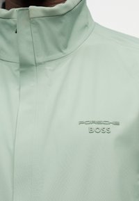 Ανοιχτό πράσινο μπουφάν με ψηλό γιακά, κρυφό φερμουάρ και ανάγλυφο λογότυπο "Porsche BOSS" στο στήθος.