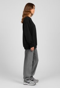 Felpa nera oversize, jeans grigi a gamba larga e sneakers grigi. Tessuto liscio, vestibilità rilassata, polsini a coste e linee pulite. Vista di profilo laterale.