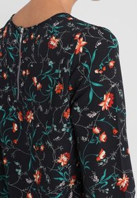 Schwarzblühende Bluse mit lebhaften roten und grünen Floralmustern, ausgestattet mit einem runden Ausschnitt und einem Reißverschlussdetail an der Rückseite. Weicher, strukturierter Stoff.