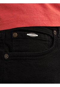 Zwarte denim jeans met benadrukte zilveren en messing hardware accenten, voorzien van een voorvak en een close-up van een merketiket.