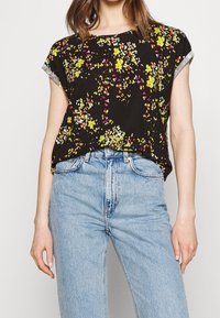 Czarny krótki top z rękawem, z kwiatowym wzorem w żółtym, różowym i zielonym kolorze. Luźny krój, okrągły dekolt i mankiety z tkaniny. Połączony z jasnoniebieskimi dżinsami.