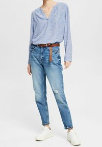 Blusa blu a fantasia con scollo a V e maniche lunghe, abbinata a jeans blu strappati e sneakers bianche. Accentuata con una cintura marrone.