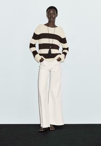 Striped sweater in beige and brown, paired with white wide-leg jeans. Accessories include a long necklace with a black tassel pendant.